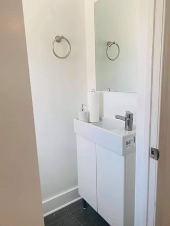 Small bright powder room with a compact white vanity sink, chrome faucet and towel ring, wall mirror, soap dispenser and paper towel roll — minimalist modern bathroom.