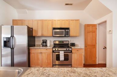 Bright modern kitchen with light wood cabinets, stainless steel side-by-side refrigerator, built-in microwave and gas range, granite countertops and coffee maker.