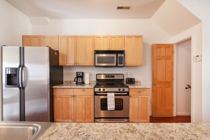 Bright modern kitchen with light wood cabinets, stainless steel side-by-side refrigerator, built-in microwave and gas range, granite countertops and coffee maker.