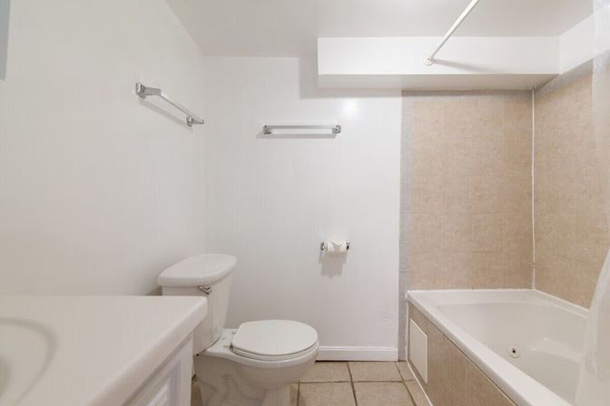 Bright minimalist white bathroom with a toilet, vanity edge, chrome towel bars, toilet paper holder, and a beige-tiled bathtub with shower rod.