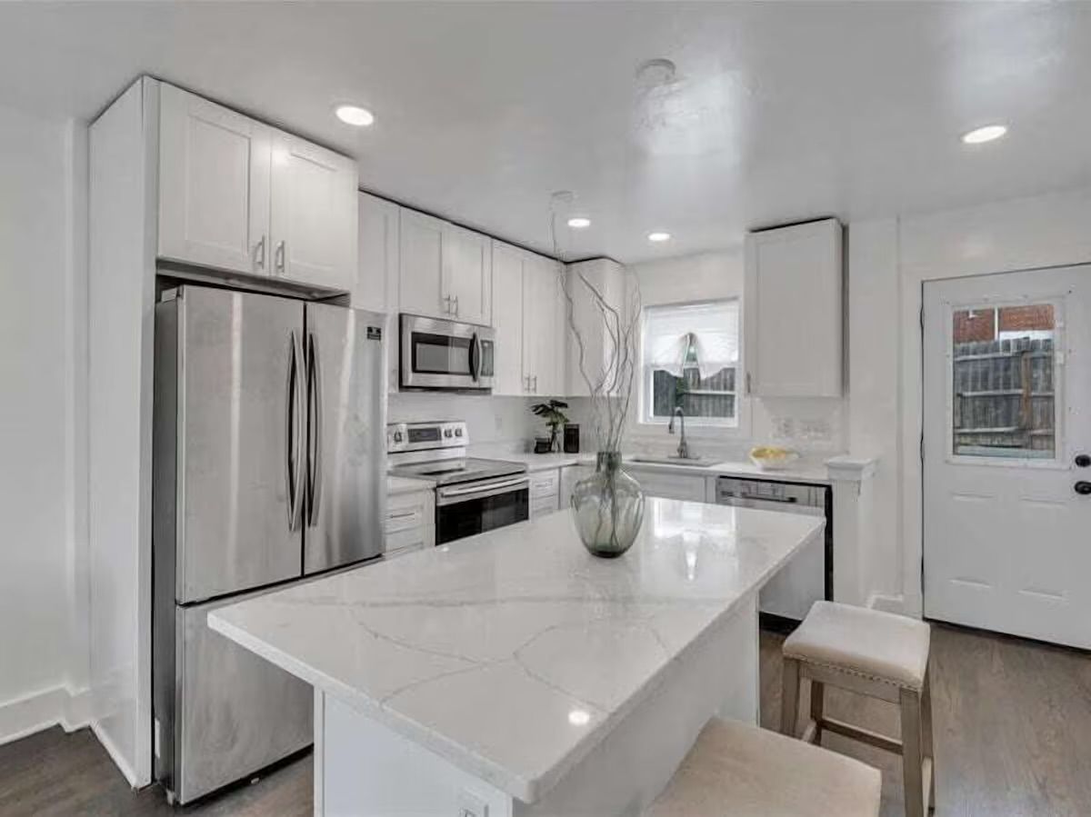 Sleek modern white kitchen with marble-look island and two stools, stainless steel refrigerator and appliances, electric range and microwave, sink under a window, recessed lighting and a back door to the yard.