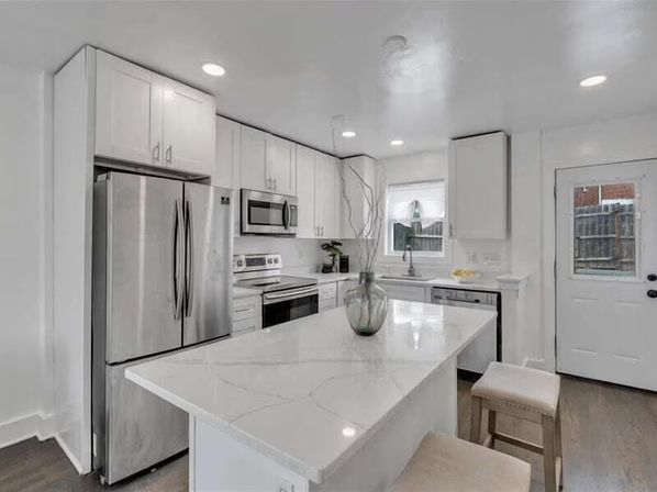 Sleek modern white kitchen with marble-look island and two stools, stainless steel refrigerator and appliances, electric range and microwave, sink under a window, recessed lighting and a back door to the yard.