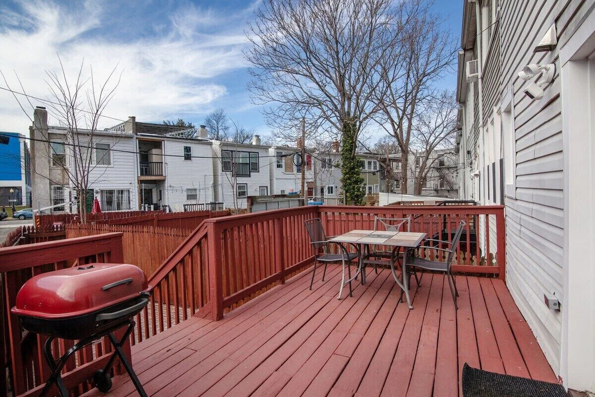 Red wooden backyard deck with a small red grill, patio table and chairs, overlooking neighboring urban row houses and leafless trees under a blue sky.