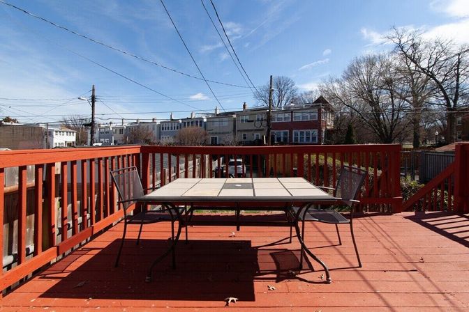 Sunny red wooden deck with a tile-top patio table and two metal chairs, overlooking urban row houses and bare trees under a bright blue sky