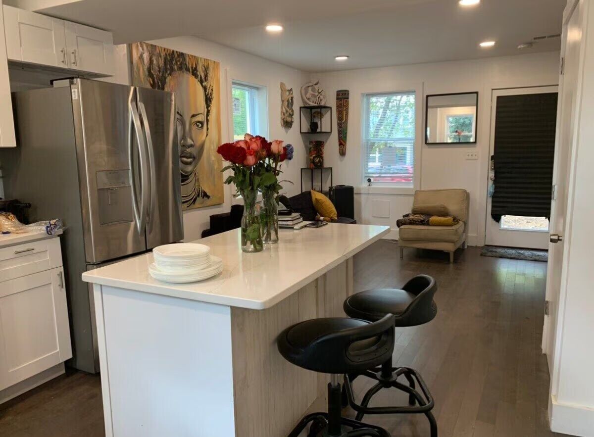 Open-concept modern kitchen and living area with white quartz island topped with a vase of red roses and stacked plates, two black swivel bar stools, stainless steel French-door refrigerator, large portrait wall art, cozy armchair by windows, and dark hardwood floors.