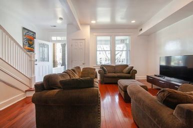 Cozy sunlit living room with red hardwood floors, plush brown sofas and ottoman, flat-screen TV, large windows and open white-trim entryway.