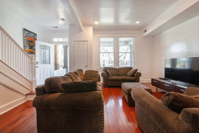 Cozy sunlit living room with red hardwood floors, plush brown sofas and ottoman, flat-screen TV, large windows and open white-trim entryway.