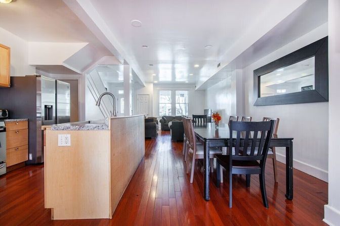 Bright open-concept kitchen and dining area with cherry hardwood floors, granite island and gooseneck faucet, stainless steel refrigerator, black dining table and chairs, large wall mirror and cozy living room by sunlit front windows.