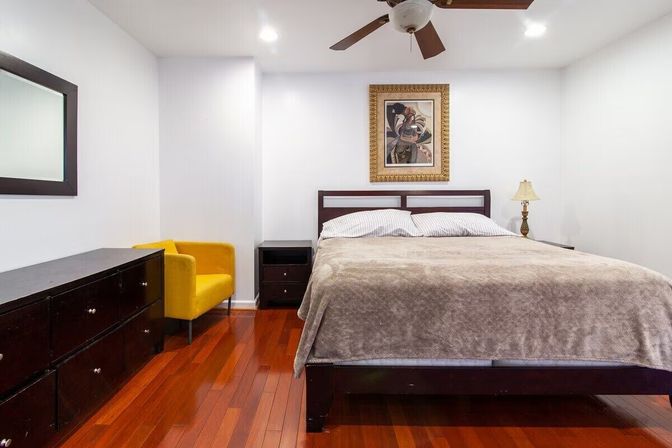 Contemporary bedroom with polished hardwood floors, large bed with neutral bedding, dark wood dresser and nightstands, yellow accent chair, framed artwork, ceiling fan and bedside lamp — bright, modern interior with a pop of color.