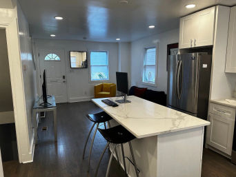 Open-concept modern kitchen and living area with white marble island and black bar stools, stainless steel fridge, yellow accent chair, computer monitor on the island, hardwood floors and recessed lighting.
