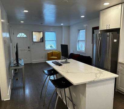 Open-concept modern kitchen and living area with white marble island and black bar stools, stainless steel fridge, yellow accent chair, computer monitor on the island, hardwood floors and recessed lighting.