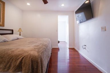 Cozy minimalist bedroom with plush beige bedspread, striped pillows, bedside lamp, ceiling fan, glossy hardwood floors and wall-mounted flat-screen TV