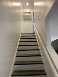 Upward view of a narrow residential stairway with white walls, dark wooden treads and a white handrail, leading to a small landing with a square mirror, a tiny access door and recessed ceiling light.