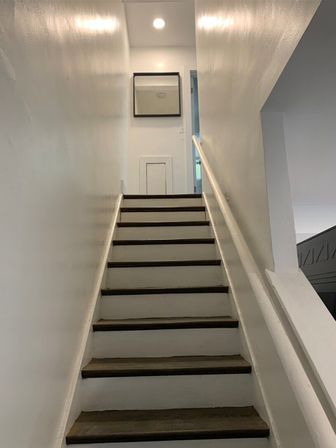 Upward view of a narrow residential stairway with white walls, dark wooden treads and a white handrail, leading to a small landing with a square mirror, a tiny access door and recessed ceiling light.