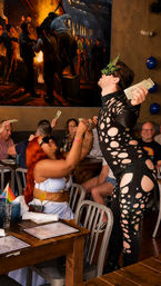 Drag performer in a black cutout rhinestone bodysuit and green mask collecting cash tips from a seated red‑haired woman at a lively indoor drag brunch, patrons cheering around a table with menus and a small pride flag.