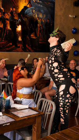 Drag performer in a black cutout rhinestone bodysuit and green mask collecting cash tips from a seated red‑haired woman at a lively indoor drag brunch, patrons cheering around a table with menus and a small pride flag.