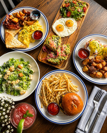 Colorful brunch spread on a wooden table: scrambled eggs with bacon, toast and home fries; sunny-side-up eggs with mixed greens; avocado toast with pickled onions; eggs Benedict with roasted potatoes; cheeseburger with fries and ketchup; Caesar salad and a pink cocktail.