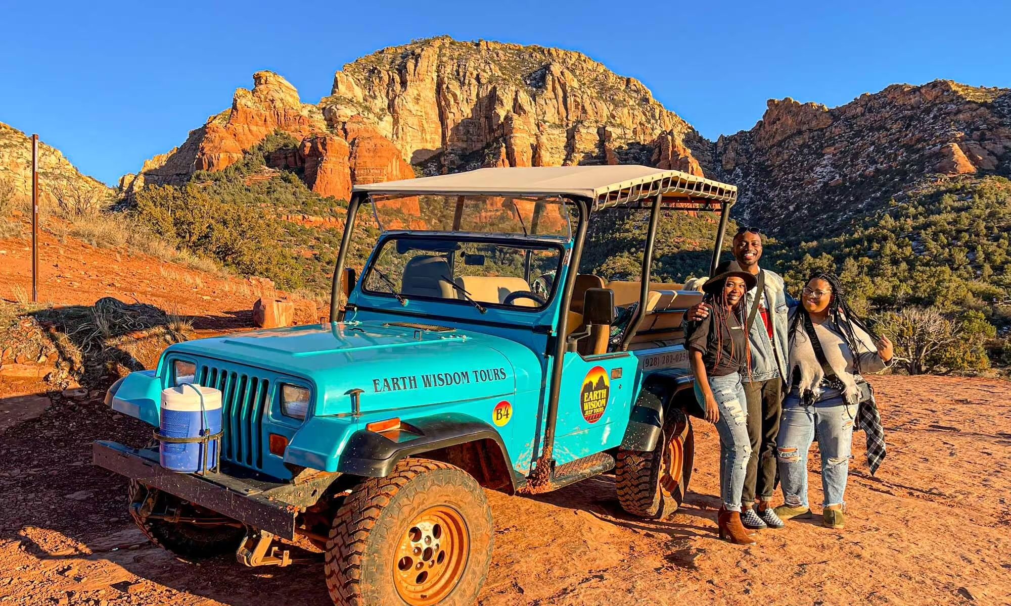 Three smiling people stand beside a bright turquoise open-air tour jeep on a red-rock trail with sunlit sandstone cliffs and a clear blue sky in Sedona, Arizona.