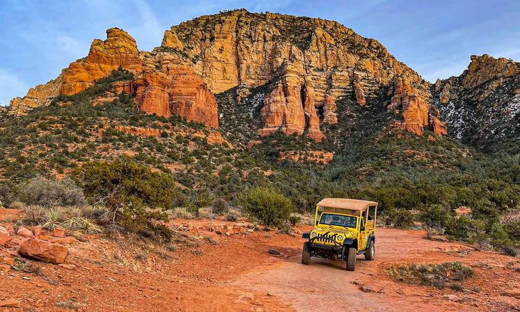 Yellow off-road jeep on a red-dirt trail in Sedona, Arizona, framed by towering red‑rock cliffs, scrubby junipers and a blue sky.