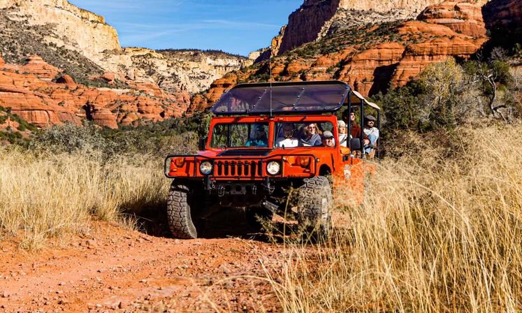 Red open-air Hummer jeep carrying tourists on an off-road tour through tall grass in a sunlit Arizona red-rock canyon under a blue sky