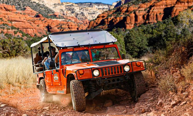 Orange open-top off-road Hummer with smiling tourists kicking up dust on a rugged red-rock trail in Sedona, Arizona, framed by sandstone cliffs and desert scrub