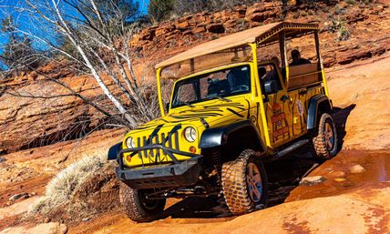 Yellow safari-style off-road jeep with black tiger-like stripes climbing a red-rock canyon trail, mud-caked tires on orange sandstone under a bright blue sky