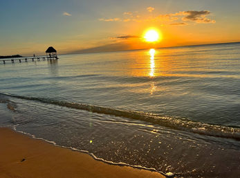 Golden tropical beach sunset with calm sea and sun reflecting on the water, gentle waves on a sandy shore, and a wooden pier with a thatched palapa silhouette.