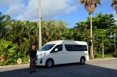 White passenger van parked on a sunlit tropical driveway lined with palm trees and blue sky, with a man in a dark shirt and sunglasses standing beside it.
