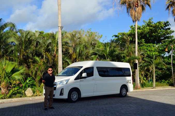 White passenger van parked on a sunlit tropical driveway lined with palm trees and blue sky, with a man in a dark shirt and sunglasses standing beside it.