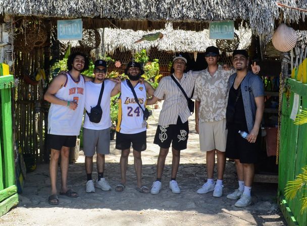 Six friends posing arm-in-arm under a thatched tropical entrance with 'Entrada' and 'Salida' signs, wearing summer outfits and basketball jerseys on a sunny vacation