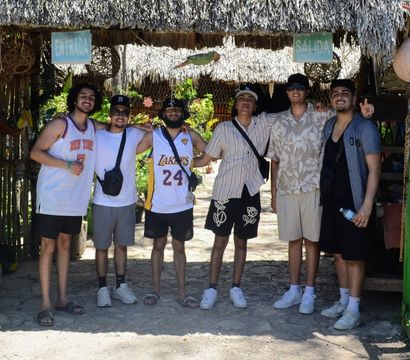 Six friends posing arm-in-arm under a thatched tropical entrance with 'Entrada' and 'Salida' signs, wearing summer outfits and basketball jerseys on a sunny vacation