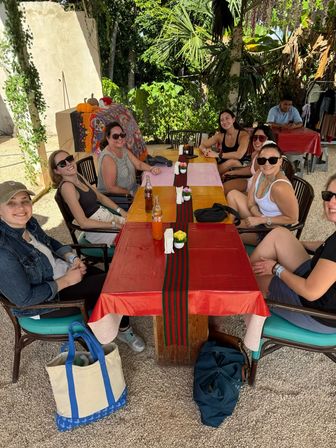 Group of friends laughing around a long wooden table at a shaded outdoor tropical patio, red and pink tablecloths with a striped runner, bottled sodas and small potted flowers, surrounded by hanging vines and palm leaves.