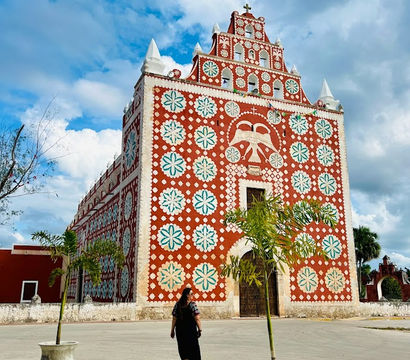 Person walking toward a tall red colonial church adorned with white-and-teal floral motifs on a sunny plaza under a vivid blue sky.