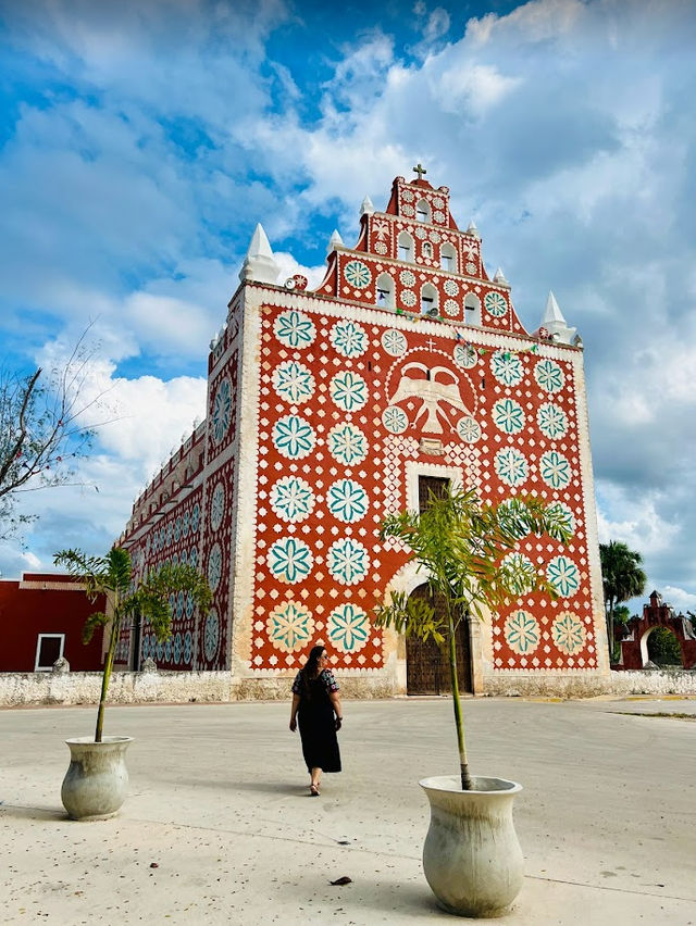 Person walking toward a tall red colonial church adorned with white-and-teal floral motifs on a sunny plaza under a vivid blue sky.