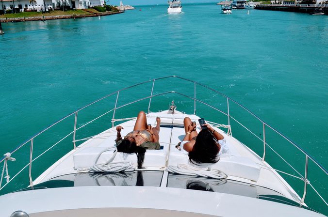 Two people lounging on the bow of a white yacht over vibrant turquoise marina waters, holding drinks and a phone with waterfront homes and moored boats in the distance.