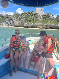 Smiling family of four in bright orange life jackets on a small boat in turquoise Caribbean water with rocky beach, palm‑topped cliff and cliffside Mayan ruins under a sunny blue sky