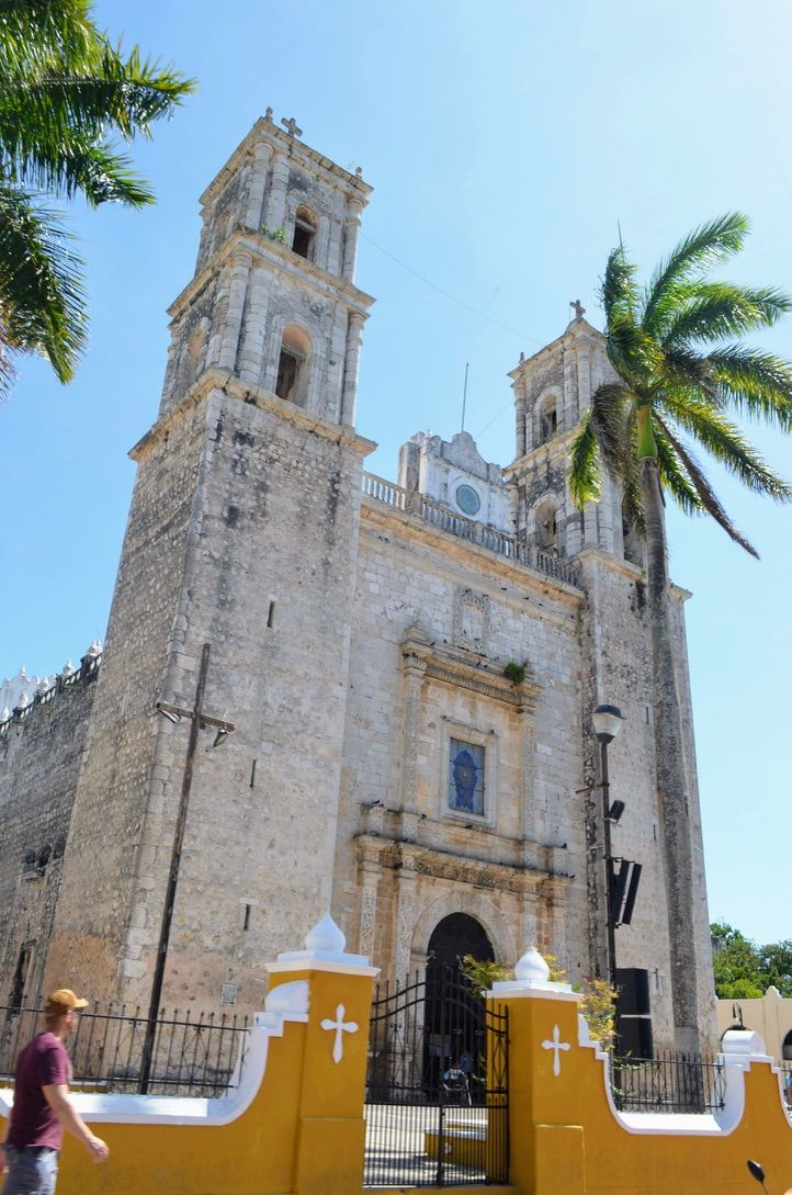Sunlit historic colonial stone church with twin bell towers, palm trees, bright yellow entrance wall and a pedestrian walking past the gate under a clear blue sky.