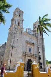 Sunlit historic colonial stone church with twin bell towers, palm trees, bright yellow entrance wall and a pedestrian walking past the gate under a clear blue sky.