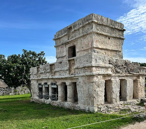 Sunlit ancient Mayan stone temple with carved façade and columned lower level on green lawn under a bright blue sky at a Yucatán coastal ruin