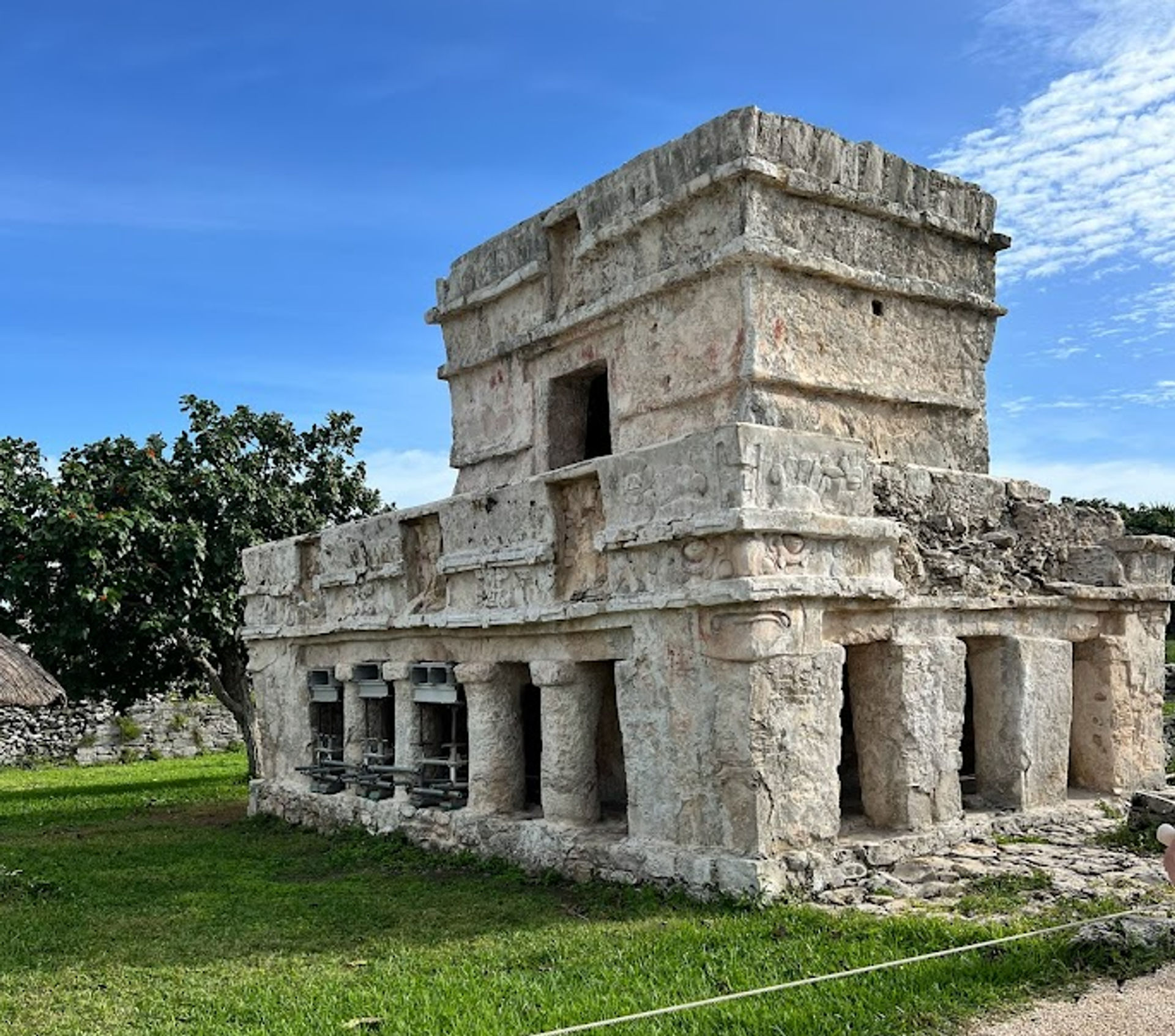 Sunlit ancient Mayan stone temple with carved façade and columned lower level on green lawn under a bright blue sky at a Yucatán coastal ruin