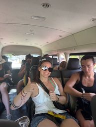 Smiling woman in sunglasses giving a thumbs-up inside a crowded tour shuttle van, surrounded by fellow travelers on a sunny vacation transfer.