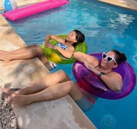 Sunny backyard pool scene with a woman in heart-shaped sunglasses and a child relaxing in colorful inflatable rings, legs propped on the pool deck for summertime float fun.
