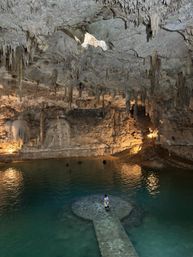 Person standing on a circular stone platform in a turquoise cenote pool inside an illuminated limestone cave, surrounded by hanging stalactites and rugged rock formations