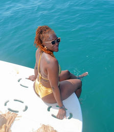 Woman in gold bikini and sunglasses sitting on a boat swim platform, dipping her feet into clear turquoise tropical sea on a sunny day
