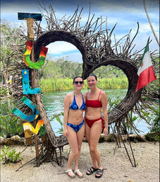 Two women in bikinis posing in front of a heart-shaped twig arch and colorful vertical 'TULUM' sign by a turquoise lagoon with a Mexican flag, Tulum, Mexico.