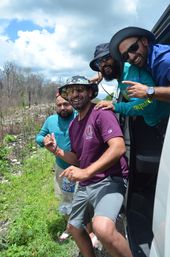Four friends leaning out of a van on a sunny rural roadside, wearing bucket hats and sunglasses, smiling and holding a water bottle.