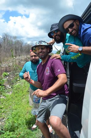 Four friends leaning out of a van on a sunny rural roadside, wearing bucket hats and sunglasses, smiling and holding a water bottle.