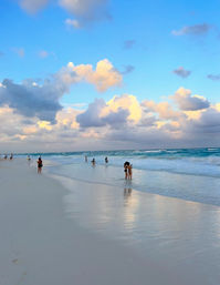 People walking and wading along a wide white-sand tropical beach with turquoise waves, fluffy golden sunset clouds and reflections on the wet shoreline.