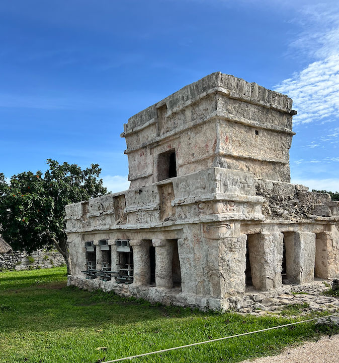 Ancient Mayan stone temple ruin with carved columns and a small upper chamber sitting on green grass under a bright blue sky