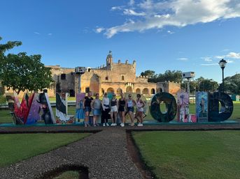 Group of visitors posing in front of colorful 'VALLADOLID' letters on a green lawn with a historic stone convent and bright blue sky in Valladolid, Yucatán, Mexico.